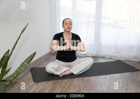 Una persona pratica yoga su un tappetino in una stanza luminosa. La persona siede a gambe incrociate con le mani unite davanti al petto. Le piante verdi sono nelle vicinanze. Foto Stock