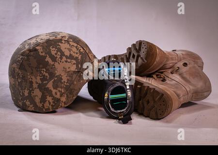Un equipaggiamento da soldato poggiato su un panno bianco. Una sistemazione tranquilla di un casco militare, stivali indossati e occhiali protettivi polverosi posizionati su tessuto bianco, simbolizina Foto Stock