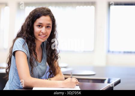 Studentessa adolescente che scrive su carta alla scrivania dell'aula tenendo la penna con una camicia azzurra. Pupilla, nottolino, luce del giorno, luce solare, luminosa, concentrati, co Foto Stock