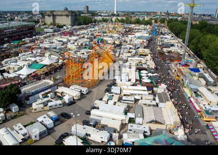 Vista aerea di una grande fiera con numerose bancarelle, giostre e attrazioni, affollate di visitatori, circondate dal verde e dagli edifici urbani Foto Stock