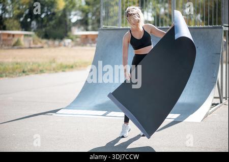 Una donna aderente all'abbigliamento sportivo regge un grande tappetino da yoga mentre si trova sul marciapiede di uno skatepark con rampe sullo sfondo Foto Stock