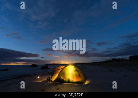 Tenda da da campeggio gialla luminosa su una spiaggia di sabbia estone sotto un cielo stellato al crepuscolo con calmo mare Baltico e pietre. Foto Stock