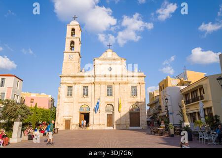 La Canee, Grecia - 21 ottobre 2022: La Cattedrale della Vergine Maria, con un campanile di primo piano e bandiere greche, si erge in una piazza illuminata dal sole con persone sedute e camminate nelle vicinanze, circondata da edifici residenziali e commerciali sotto un cielo parzialmente nuvoloso. Foto Stock