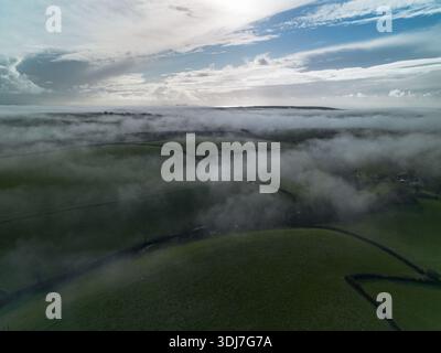 Vista aerea di nuvole basse e nebbie che si muovono attraverso i verdi terreni agricoli e i siepi sotto un cielo nuvoloso spettacolare. Suggestivo paesaggio rurale. Foto Stock