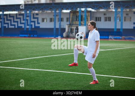 Giocatrice di calcio in uniforme bianca che giocherà con palla durante le esercitazioni sul campo verde in uno stadio all'aperto. Foto Stock