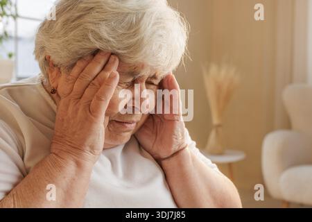 Primo piano di una donna anziana che tocca i templi, soffre di mal di testa o emicrania a casa, occhi chiusi nel dolore. Foto Stock