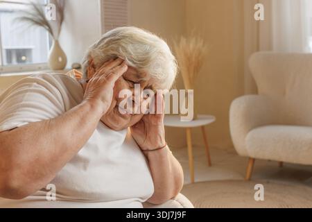 Donna anziana che soffre di mal di testa, che tiene i templi nel dolore mentre è seduta a casa, sfondo con luce naturale. Foto Stock