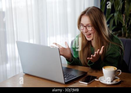 Giovane donna eccitata con gli occhiali che reagisce con gioia mentre videochiama con un laptop in un bar con caffè. Foto Stock