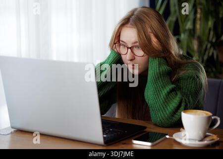 Giovane donna frustrata che guarda lo schermo di un notebook mentre lavora in remoto in un bar, affrontando problemi tecnici o lavorativi. Foto Stock