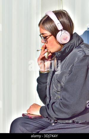Giovane donna/adolescente che indossa le cuffie, fuma e tiene in mano lo smartphone - Loches, Indre-et-Loire (37), Francia. Foto Stock