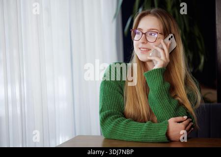 Sorridente giovane donna in occhiali che parla sullo smartphone mentre siede a un tavolo accanto alla finestra in un accogliente caffè. Foto Stock