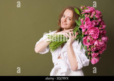 Grande bouquet di peonie rosa tenuto da una donna in bianco su sfondo verde oliva. Foto Stock