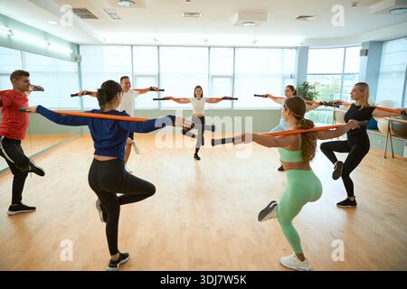 Gruppo di persone che posa in cerchio in equilibrio sulla gamba tenendo i bastoncini da ginnastica sulle spalle Foto Stock