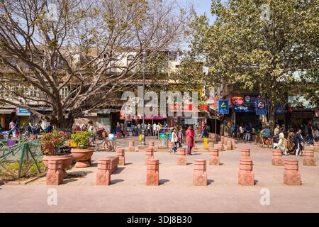 Delhi, India - 13 marzo 2022: Le persone camminano e si fermano vicino a grandi alberi, dissuasori di pietra rossa e segnaletica commerciale di fronte a negozi e vecchi edifici a più piani lungo una strada nella vecchia Delhi. Foto Stock