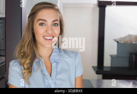 Seduto alla reception, donna adulta che indossa una camicetta blu chiaro sorridente e che ordina scatole grigie. Professionale, ufficio, organizzato, tranquillo, efficiente, admi Foto Stock
