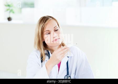 Medico donna che esamina il termometro in una stanza clinica luminosa con camice da laboratorio bianco e stetoscopio. Clinico, sanitario, medico, esame, calma, Foto Stock