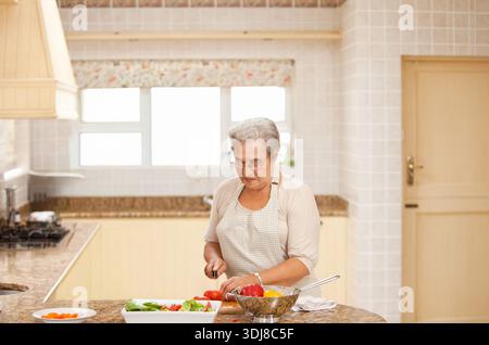 Donna anziana che trita pomodori e mescola insalata sull'isola di granito con grembiule a scacchi e coltello. Cucina, soleggiata, accogliente, casalinga, verdure, sl Foto Stock
