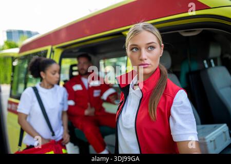 Tre paramedici multirazziali in piedi davanti al veicolo dell'ambulanza, che trasportano apparecchiature portatili Foto Stock