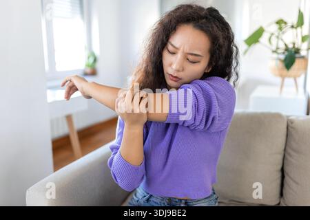 infelice donna asiatica con dolore al gomito al chiuso. sindrome da ufficio concetto di assistenza sanitaria. sconvolta ragazza asiatica disorientata confuso guardando le braccia ferendo seduto o Foto Stock
