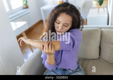 infelice donna asiatica con dolore al gomito al chiuso. sindrome da ufficio concetto di assistenza sanitaria. sconvolta ragazza asiatica disorientata confuso guardando le braccia ferendo seduto o Foto Stock