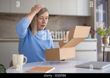 Una donna anziana a casa è rimasta delusa dopo aver aperto un ordine online, tenendo una scatola di cartone vuota e danneggiata al tavolo della cucina, frustrata per l'articolo sbagliato e il servizio di consegna scarso Foto Stock