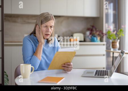 Una donna anziana seduta al tavolo della cucina con un portatile, che legge un conto angosciante in una busta aperta, visibilmente preoccupata e sopraffatta da notizie finanziarie inaspettate Foto Stock