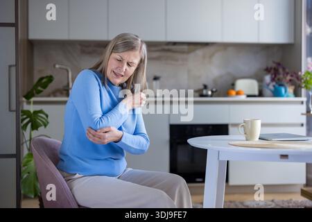 Donna anziana seduta a un tavolo da cucina moderno, che si aggrappa e stringe il gomito con entrambe le mani, soffre di dolori articolari e disagio per artrite o lesioni a casa Foto Stock