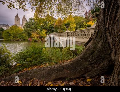 Bow Bridge a Central Park incorniciato da un fogliame autunnale, con le torri gemelle di San Remo che si innalzano sopra Central Park West a Manhattan, New York. Foto Stock