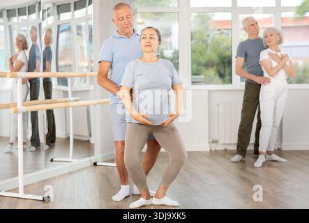 Diligente coppia di ballerini moderni anziani che praticano il balletto demi plie con supporto nella sala con bar di balletto Foto Stock