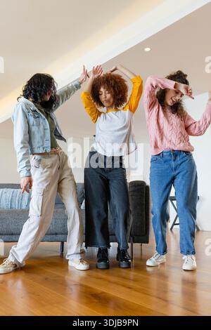 Tre amiche che ballano in salotto su un pavimento in legno duro su un divano grigio con scarpe da ginnastica Foto Stock