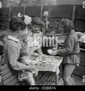 Ucraina, regione di Donetsk, Sloviansk. Circa 1960 anni Una foto d'archivio in bianco e nero che mostra un gruppo di bambini che giocano alla festa del tè in un parco giochi dell'asilo. La ragazza a sinistra con un maglione a motivi che tiene in mano una bambola è Inessa Mikhailovna Kravelis. Gli amici sistemano i piatti dei giocattoli su un tavolino ricoperto di un panno. Ragazzi e ragazze sono impegnati in un gioco di ruolo con un set da tè. La scena cattura la socializzazione e il processo educativo dei bambini in età prescolare. L'atmosfera di un'infanzia felice e spensierata in URSS. Una vista storica del tranquillo Donbas. Foto Stock