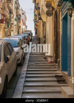 La Valletta, Malta - 8 aprile 2024: Una stretta strada a gradini a la Valletta fiancheggiata da auto parcheggiate e tradizionali edifici calcarei, che illustrano il cit Foto Stock