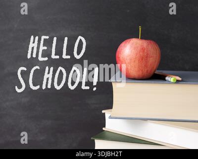 Una pila di libri, mela rossa e matita con un messaggio di saluto scritto in gesso su una lavagna, concetto di ritorno a scuola e istruzione Foto Stock
