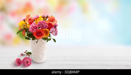 Un vivace bouquet di rose e altri fiori primaverili in sfumature di rosa, rosso, arancione e giallo è disposto in un vaso bianco ornato. Il vaso si trova su un Foto Stock