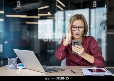 Giovane donna d'affari stressata che reagisce a cattive notizie o a problemi imprevisti sul suo smartphone, è scioccata e frustrata mentre lavora alla sua scrivania in un ufficio moderno Foto Stock