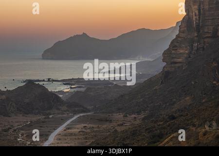 Un'ampia vista aerea della spettacolare costa dell'Oman al tramonto. Una strada tortuosa serpeggia lungo la riva, fiancheggiata da aspre montagne e dalla tranquilla A Foto Stock