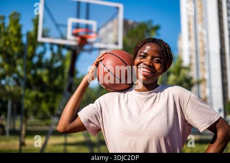 Giovane donna afroamericana sorridente mentre tiene il basket in campo urbano all'aperto. Foto Stock