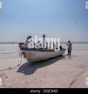 Un gruppo di pescatori dell'Oman su una spiaggia sabbiosa che si prepara o torna dalla pesca, con una tradizionale barca da pesca bianca e blu in primo piano. Foto Stock