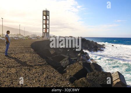 Puerto de la Cruz, 01152026 - uomo in piedi sulla costa rocciosa vicino al faro e Breakwater con onde che si schiantano Foto Stock