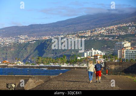 Puerto de la Cruz, 01152026 - coppia che cammina lungo la passeggiata costiera con paesaggio urbano e vista sull'oceano Foto Stock