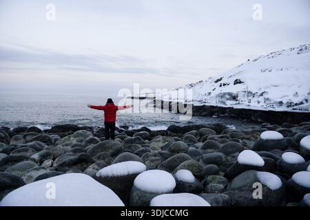 Vista posteriore di un uomo con uno stemma di pompieri in piedi con le braccia allungate sulla costa del Mare di Barents in inverno, Teriberka, distretto di Kolsky, Oblast di Murmansk, Russia Foto Stock