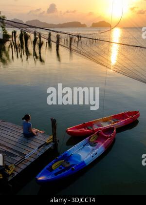 Vista dall'alto di una donna seduta su un molo di legno con due kayak ormeggiati accanto al tramonto, Ko Mat, lago Songkhla, Phatthalung, Thailandia Foto Stock
