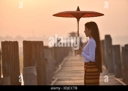 Vista laterale di una giovane donna che porta un ombrellone tradizionale in piedi sul ponte U Bein, Amarapura vicino a Mandalay, Myanmar Foto Stock