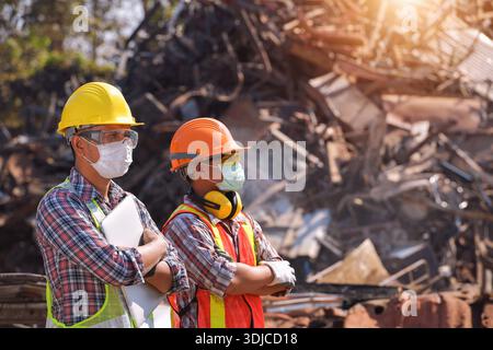 Due lavoratori manuali che indossano maschere facciali e indumenti da lavoro protettivi in piedi fianco a fianco in un deposito di rottami di metallo, Thailandia Foto Stock