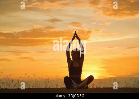 Silhouette di una donna seduta a gambe incrociate con le braccia in aria che fa yoga al tramonto, Thailandia Foto Stock