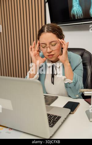 Donna che sente dolore nei templi mentre è seduta alla scrivania con un notebook Foto Stock