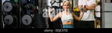 Intestazione di donna caucasica di mezza età che esegue esercizio alla spalla con manubri, sotto la supervisione di un uomo caucasico di mezza età, in palestra durante la sessione di riabilitazione di terapia fisica Foto Stock