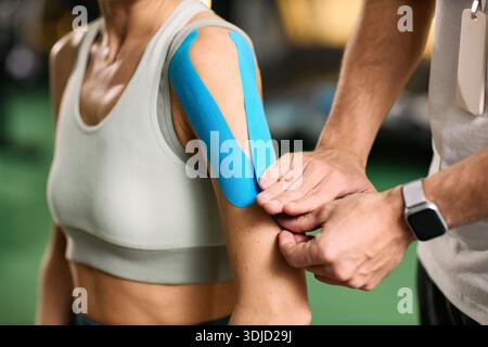 Donna caucasica di mezza età che riceve l'applicazione del nastro per kinesiologia sulla parte superiore del braccio da un fisioterapista maschio durante una sessione di riabilitazione in ambiente clinico Foto Stock