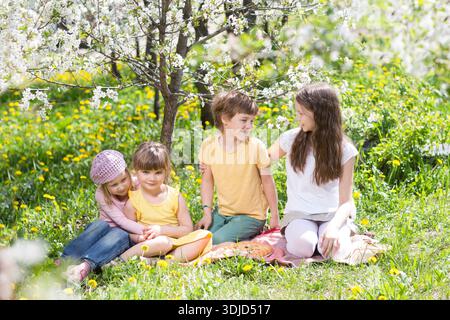 i bambini e gli amici trascorrono del tempo insieme nella natura. Un gruppo di bambini si siede sull'erba e fa un picnic nel giardino di primavera || modello rilasciato Foto Stock