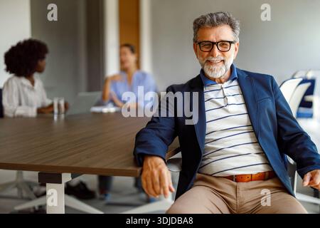 Ritratto di un manager maschio in occhiali che sorride con sicurezza mentre siede in sala riunioni con colleghi che collaborano sullo sfondo Foto Stock